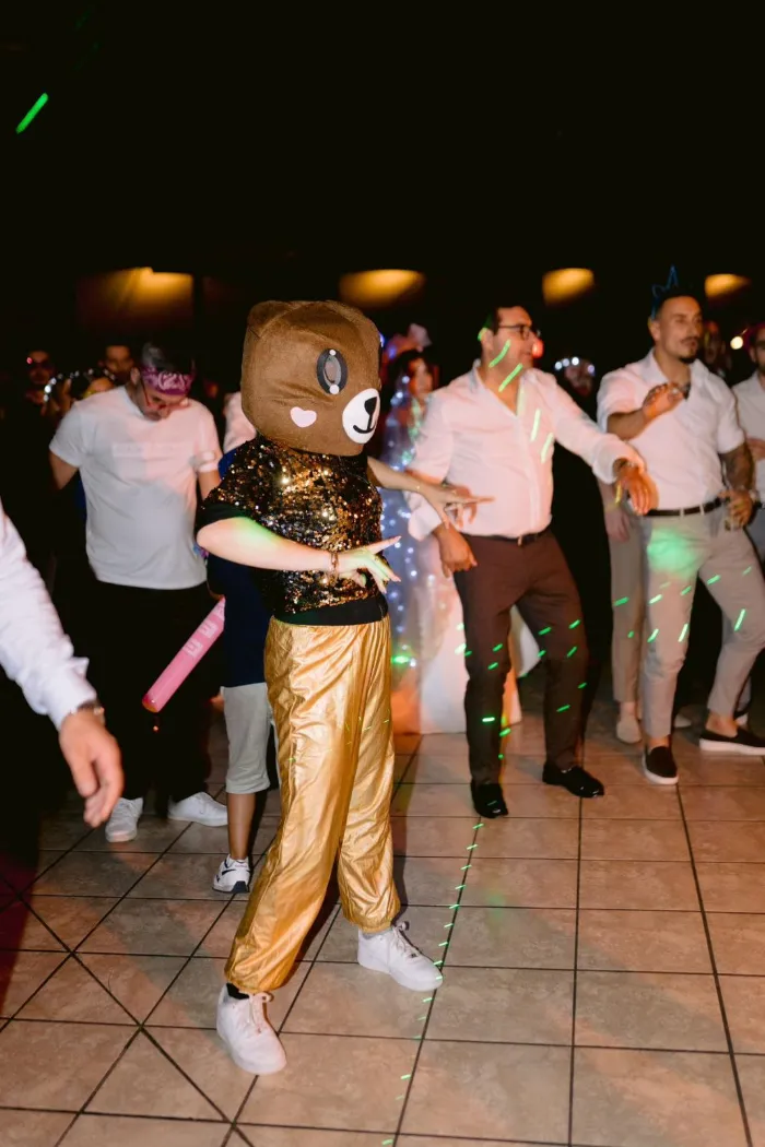 Mascote urso da PlayDay, de calças douradas, a dançar na pista no meio dos convidados de um casamento.